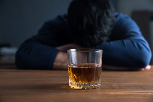 glass of alcohol on table while man sleeps on his arms on table wondering is alcoholism is disease