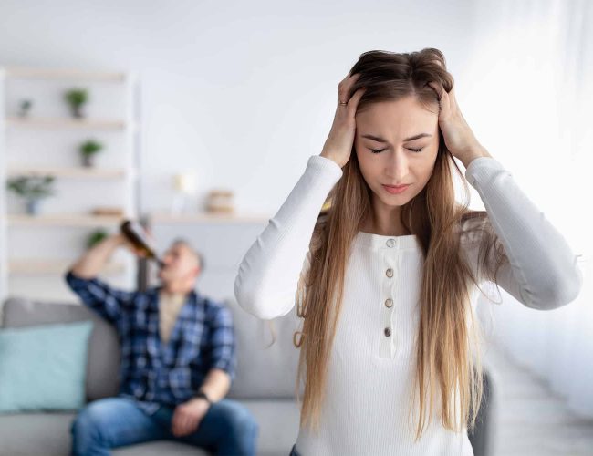 woman holding her head in frustration with her husbad sitting on the couch behind her drinking alcohol
