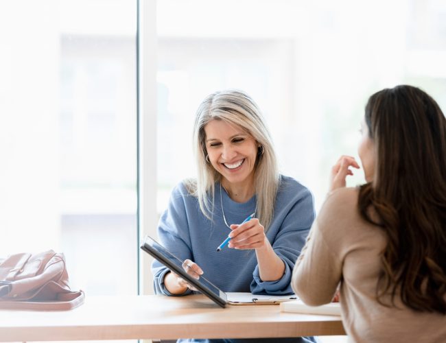 Meeting with the unrecognizable female client, the mature adult female financial advisor goes over documents on the digital tablet.