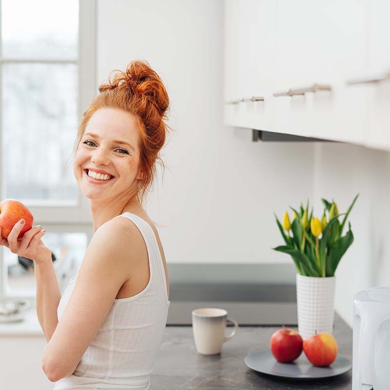 woman standing in the kitchen eating an apple for her nutritional therapy program