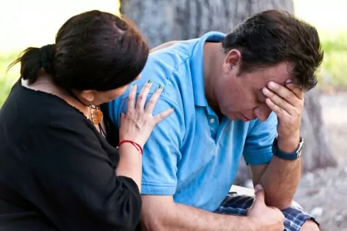 Man bent over holding his head while a woman embraces him