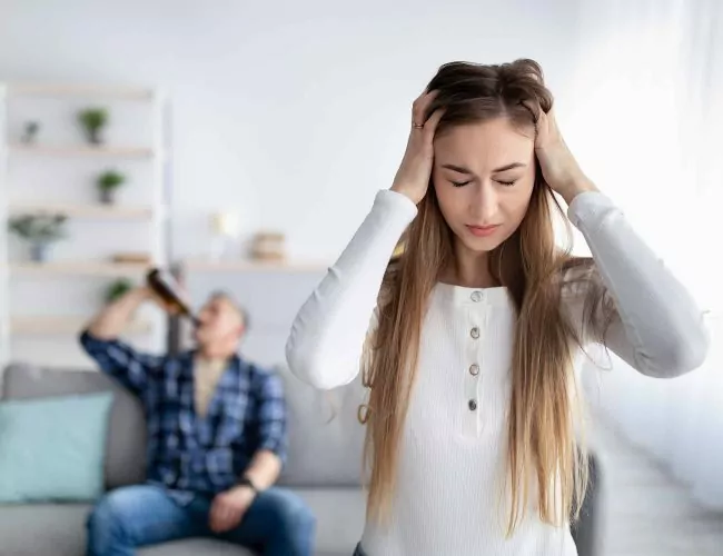 woman holding her head in frustration with her husbad sitting on the couch behind her drinking alcohol